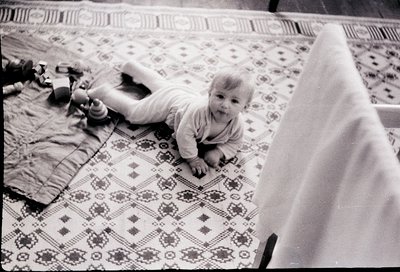 Black-and-white snapshot of a toddler crawling on a patterned rug in a mid-century home. Distinctive floral motifs and geomet...
