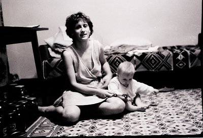 A woman and toddler share a candid moment in a mid-20th-century home. The woman, seated on patterned rugs, holds a toy boat, ...