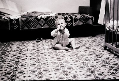 Black-and-white snapshot of an infant crawling on patterned rug, holding a pacifier. Geometric textile designs on bedding and...