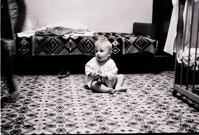 Mid-20th century black-and-white photo of a toddler sitting on patterned rug, playing with a toy. Geometric bedspread with di...