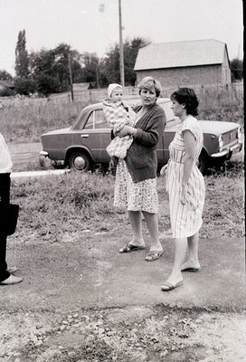 Mid-20th century rural scene featuring three women: one holding a baby in a floral dress, another in a striped dress carrying...