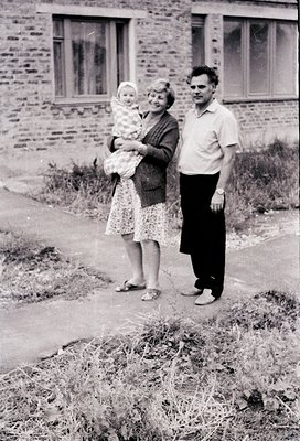 Family portrait in front of a brick residential building, likely Eastern Bloc-era (1960s–1970s). Woman holds a baby in a patt...