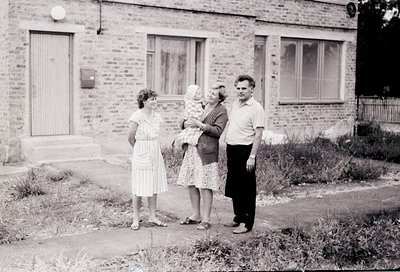 Four individuals pose outside a brick residential building, likely mid-20th century. The woman on the left wears a fitted dre...