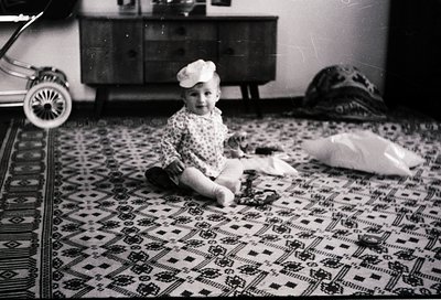 Vintage black-and-white photo of a toddler sitting on an intricately patterned rug in a mid-century home. The child wears a p...