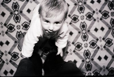 Vintage black-and-white photo of a child making a peace sign against a patterned wallpaper backdrop. Mid-20th century indoor ...