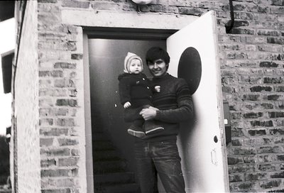 A man in mid-20th-century attire holds a baby at a brick entranceway, framed by a white door. The man wears a dark jacket wit...