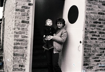 A woman holds a toddler in a brick-lined hallway, framed by a white door with a black circular design. Mid-20th century urban...