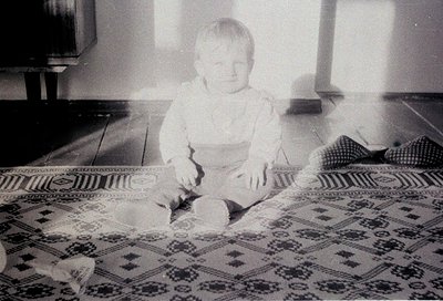 Vintage black-and-white photo of a toddler sitting on a patterned rug with geometric designs, holding a small object. Mid-cen...