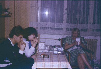 Three individuals seated indoors in a dimly lit room, likely mid-1970s–1980s Eastern Bloc. Man in suit (left) holds a badge, ...