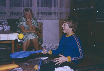 Vintage indoor ping-pong scene featuring two people in 1970s attire. Woman in floral dress observes while man in blue sweater...