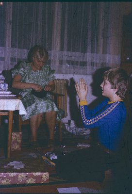 Vintage indoor scene featuring an elderly woman in floral dress sewing while a young boy in a blue embroidered shirt kneels b...