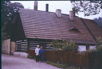 Two individuals pose outside a rustic log cabin-style house with a steep, shingled roof and chimneys, framed by a wooden pick...