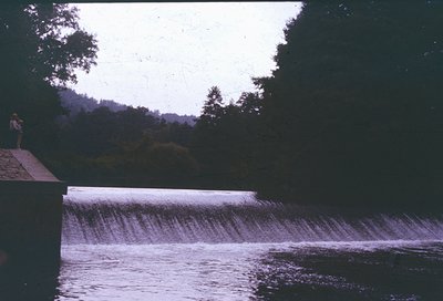 Vintage photo of a concrete dam spillway with cascading water under overcast skies. A lone figure stands on a concrete ledge,...