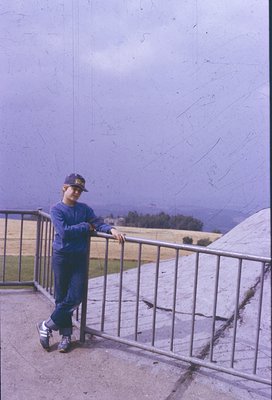 Vintage photo of a young boy in a blue cap and matching tracksuit posing on a concrete balcony overlooking a misty, hilly lan...
