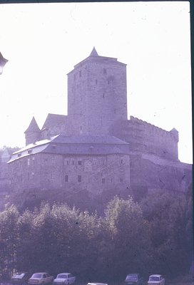 Medieval stone fortress with cylindrical tower and crenellated walls, likely European. Foreground shows vintage cars and tree...