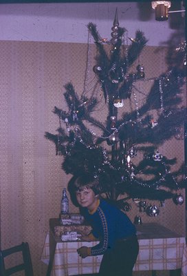 Vintage Christmas scene featuring a young girl seated beside a modestly decorated pine tree adorned with tinsel, baubles, and...