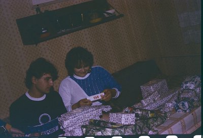 Vintage indoor scene featuring three individuals playing cards on a bed with floral-patterned bedding. The man on left wears ...