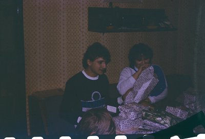 Vintage indoor scene featuring three individuals seated at a patterned table, likely from the 1970s–1980s. The man wears a da...