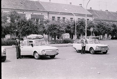 Vintage street scene featuring two Eastern Bloc-era cars (likely Škoda 100/110/120) parked on an urban road. One car has a la...