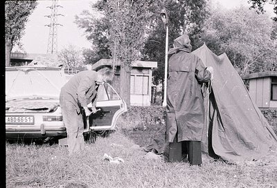 Black-and-white street scene featuring two individuals assisting a woman into a vintage car (ABD 66-51) near a makeshift tent...