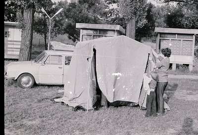 A 1960s-era camping scene: a woman and child setting up a canvas tent beside a vintage car in a grassy area. Nearby, a bus sh...