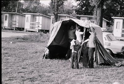 Mid-20th century family camping scene in a grassy area with mobile homes in background. Three adults and a child assist setti...