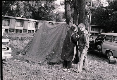 Black-and-white photo of two individuals setting up a canvas tent in a grassy area, likely mid-1960s–1970s. Residential build...