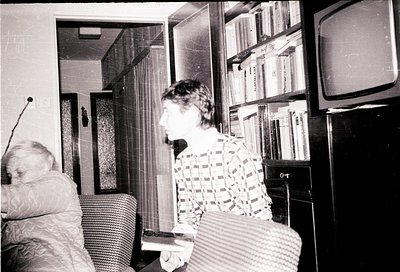 Vintage black-and-white interior shot of a mid-century living room. A woman in a patterned blouse sits on a woven armchair, h...