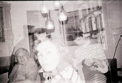 Vintage black-and-white photo of two women in a dimly lit indoor setting, likely mid-20th century. One woman sits on a chair,...
