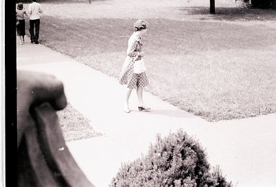 Mid-20th century street scene: woman in checkered dress walks past mannequin arm on sidewalk, blurred foreground. Grass lawn ...