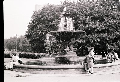 Classic mid-20th-century fountain with sculpted sea creatures, surrounded by lush greenery. People in vintage attire—men in s...