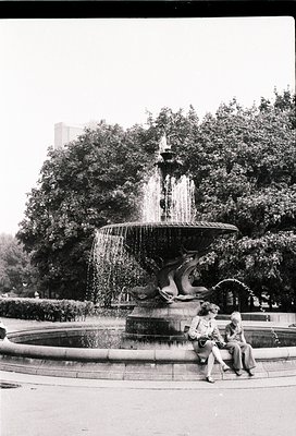 Vintage black-and-white shot of a multi-tiered fountain with sculpted sea creatures, likely from the 1950s–1960s. Two seated ...