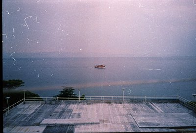 Vintage seaside view through a slightly scratched window, showing a lone boat on calm waters. Concrete balcony with metal rai...