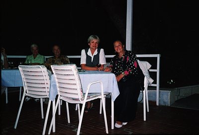 Four adults seated at an outdoor table in a dimly lit, open-air dining area. White plastic chairs and tablecloths suggest a m...