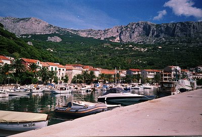 Coastal marina nestled between rugged mountains and Mediterranean-style buildings. Small boats docked along calm waters, with...