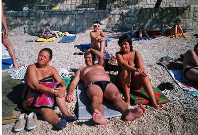 Three women relax on a pebble beach, seated on towels under bright sunlight. The central figure wears a swimsuit with a patte...
