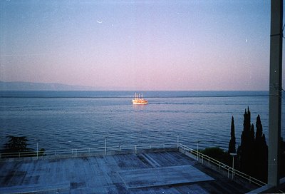 Vintage seaside balcony view with a lone sailboat on calm waters at dusk. Concrete railing and potted cypress trees frame the...