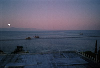 Twilight seascape with three small wooden boats anchored near shore. Concrete terrace with metal railings in foreground. Dist...