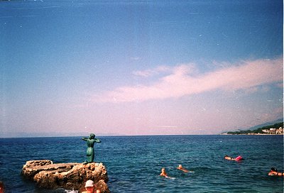 Aerial view of a shallow, sandy beach with clear turquoise waters. Three swimmers in the background near rocky formations. Vi...