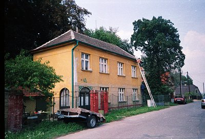 Two-story yellow building with red-tiled roof, likely residential or small commercial, featuring arched entrance and white-fr...