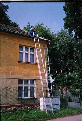 Aerial view of a mid-rise building under maintenance, featuring a white extension ladder against a yellowish stucco facade. T...