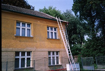 Two-story yellow stucco building with white-framed windows, likely mid-20th century. Ladder leans against roof, suggesting ma...