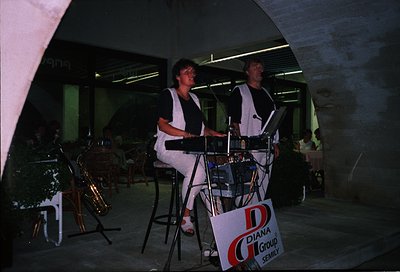 Two musicians performing live in a dimly lit, modernist indoor venue. The woman plays a keyboard, the man sings into a microp...