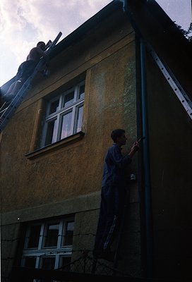 A rooftop view of a weathered concrete building facade, showing aged yellowish plaster and exposed rebar. A lone person in da...
