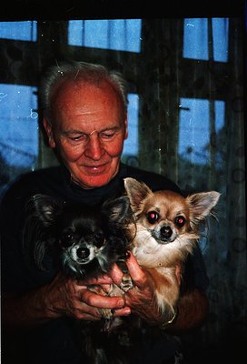 Vintage portrait of an elderly man holding two small dogs (likely Pomeranians) indoors, framed by a blue-tinted window. Warm ...