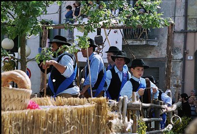 Traditional rural procession with men and boys in matching blue-and-white folk attire, carrying agricultural tools and bundle...