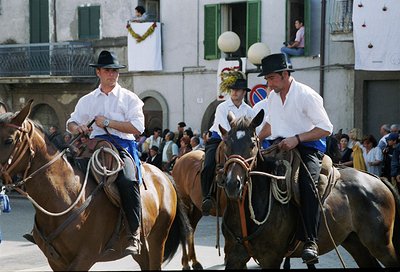 Riders in traditional white shirts, black hats, and blue trousers lead horses in a ceremonial procession through a European t...