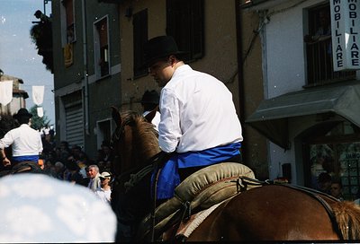 Rider in traditional alpine attire (white shirt, blue sash, black cap) leads a brown horse through a crowded street. Surround...
