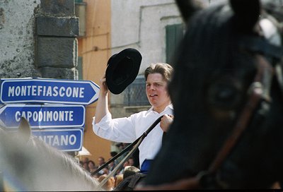 A man in a white shirt holds a fedora aloft near blue directional road signs ("Montefiascone," "Capodimonte," "Sant'Anna") in...