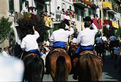 Three horsemen in traditional white jackets, blue sashes, and black hats carry floral arrangements during a festive processio...